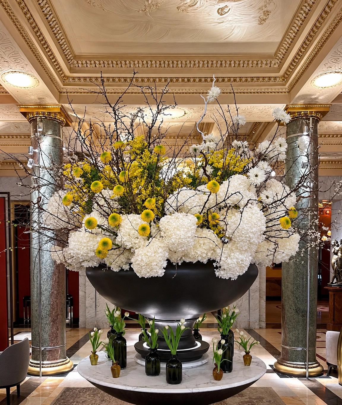 White hydrangeas, yellow flowers, and airy branches in a large dark lobby bowl